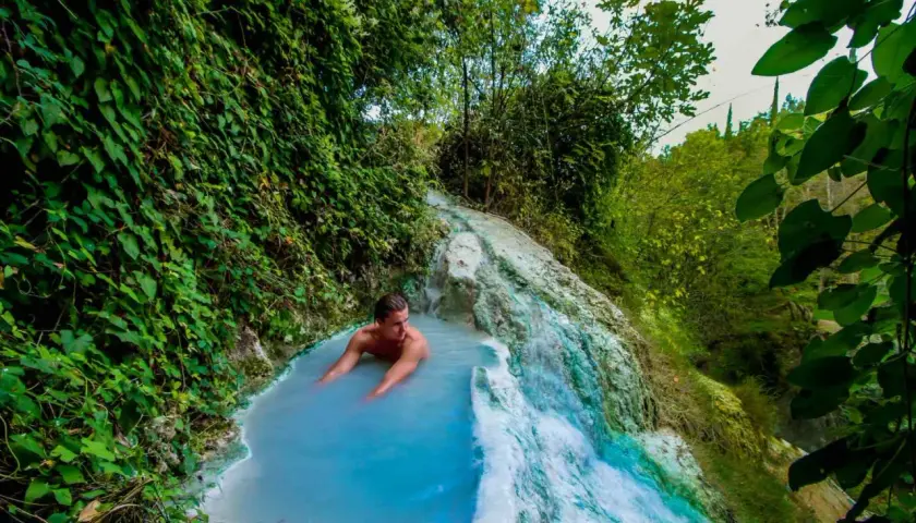 Un uomo si rilassa nelle acque azzurre di una cascata termale naturale a Olmitello, Ischia, circondato da lussureggiante vegetazione.