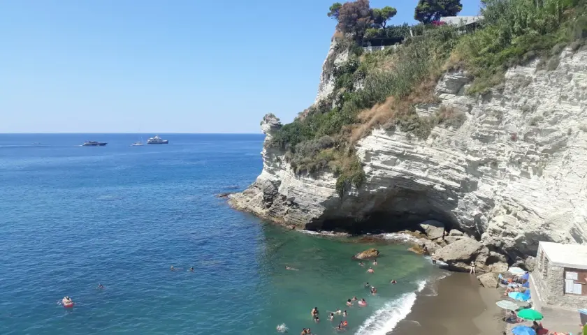 Scogliera bianca a picco sul mare turchese, con bagnanti e yacht all'orizzonte. La **cava grado Ischia** offre un rifugio naturale.