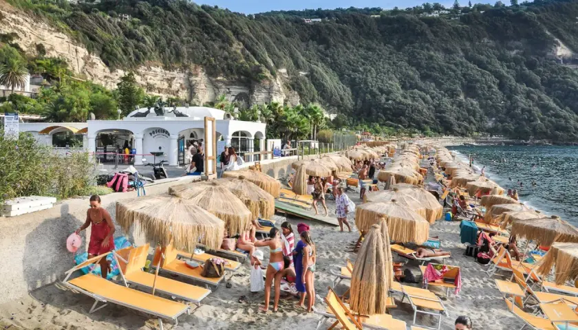 Spiaggia di Citara affollata, con ombrelloni di paglia, lettini arancioni e gente che si gode il mare. Per arrivare, segui le indicazioni per Forio.