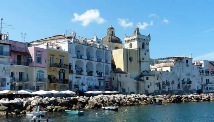 Colorate case affacciate sul mare, un porto con barche e rocce. Per sapere come arrivare ad Ischia in treno, si può prendere un treno per Napoli e poi un aliscafo.
