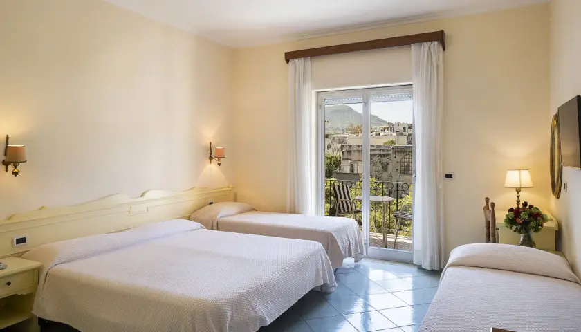Camera con tre letti singoli, vista su un borgo di Alexander Ischia. Balcone con tavolo e sedie.