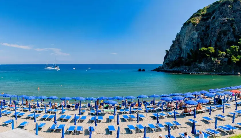 Spiaggia affollata con ombrelloni blu e lettini sulla riviera di San Montano. Mare azzurro e scogliera verde sullo sfondo.