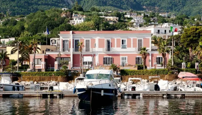 Il **hotel Svizzera Ischia** si affaccia sul porto con barche ormeggiate. Sullo sfondo, case arroccate su una collina verdeggiante.