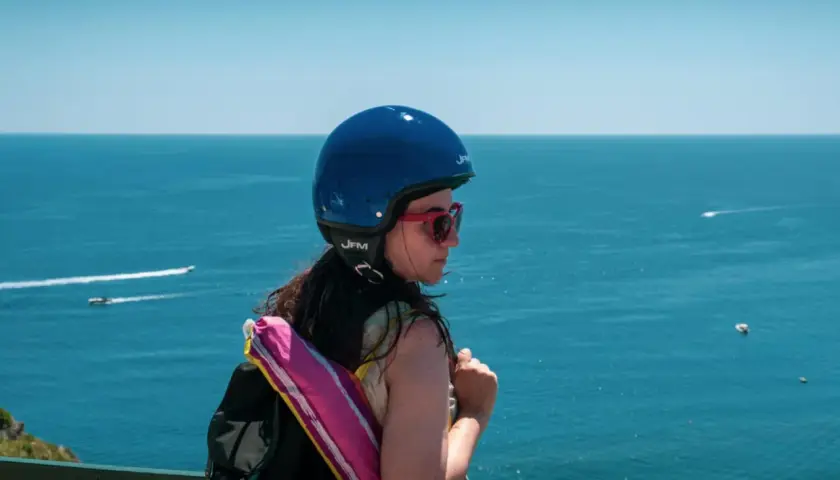 Ragazza con casco blu e zaino, guarda il mare. Pensa a come arrivare ad Ischia in auto, magari con uno scooter.