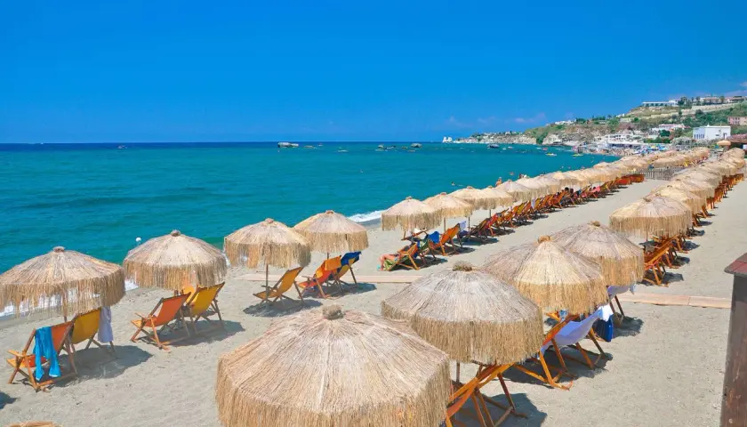Spiaggia attrezzata con ombrelloni e lettini, mare azzurro e case in collina. Perfetto per chi cerca il **hotel San Valentino Ischia distanza dal mare** ideale.