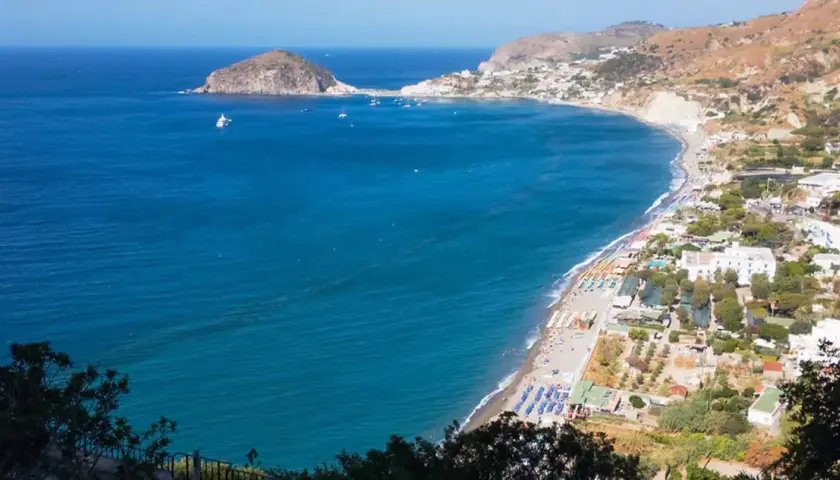 Vista panoramica della spiaggia dei Maronti con lidi attrezzati, ombrelloni colorati e il mare blu intenso. In lontananza, un isolotto e la costa frastagliata.