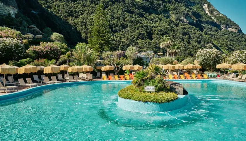 Piscina con lettini e ombrelloni a Ischia, immersa nel verde rigoglioso. Un'oasi di relax che ricorda la forza del **Tifeo** che dorme sotto l'isola.
