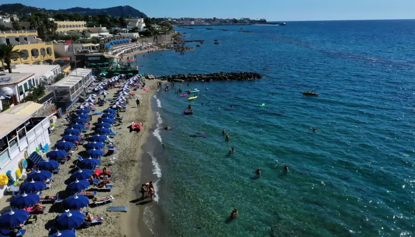 Forio Ischia spiagge affollate sotto il sole. Ombrelloni blu, bagnanti in mare e hotel affacciati sulla costa.
