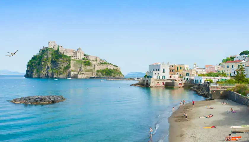 Lidi di Ischia Porto con il Castello Aragonese sullo sfondo. Gente in spiaggia e mare azzurro sotto un cielo limpido.