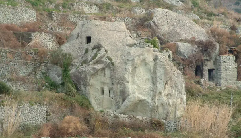 Antiche case in pietra scavate nella roccia e costruite con muretti a secco, immerse in un paesaggio collinare brullo.