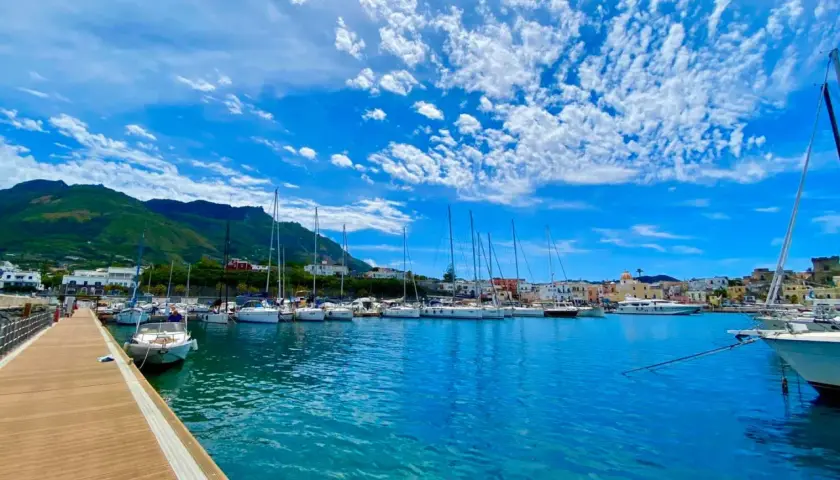 Porto di Forio, Ischia: barche a vela ormeggiate in un mare azzurro sotto un cielo con nuvole bianche. Per raggiungere Forio da Ischia Porto, si può prendere un autobus o un taxi.