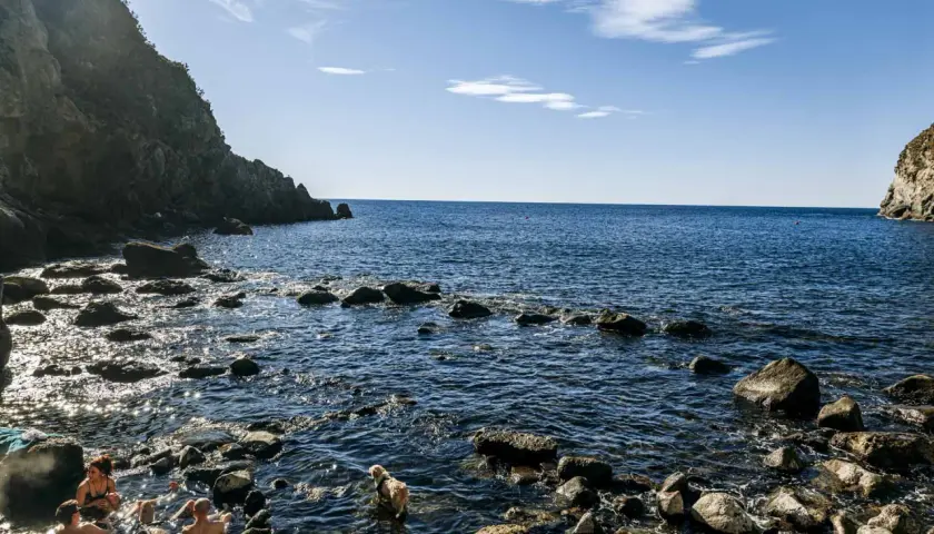 Persone e un cane si godono le acque calde della **baia di Sorgeto**, tra scogli e rocce a picco sul mare blu.