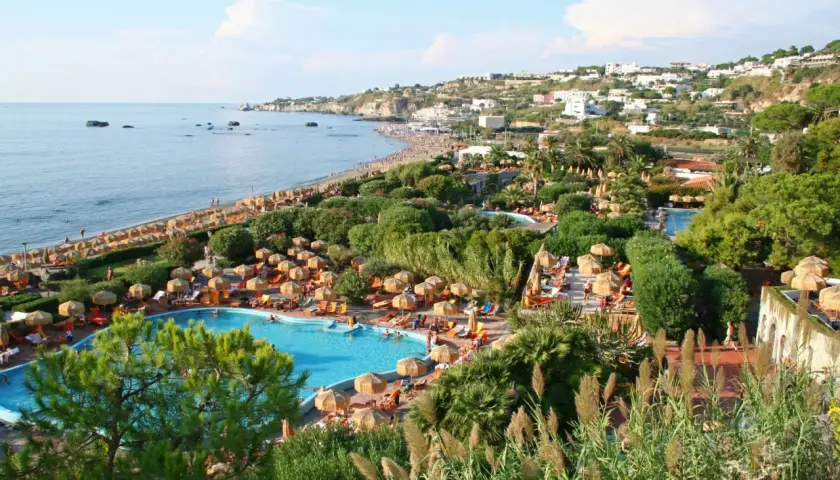 Vista panoramica di un resort a Ischia con piscine, lettini, ombrelloni e la spiaggia che si estende verso il mare.