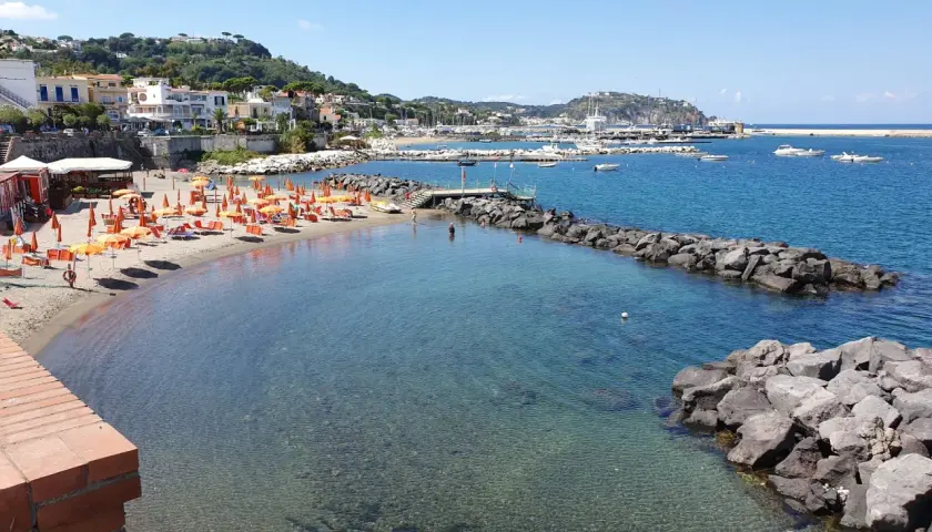 La spiaggia della marina è baciata dal sole, con ombrelloni arancioni e lettini pronti per accogliere i bagnanti. Il mare cristallino invita a un tuffo.