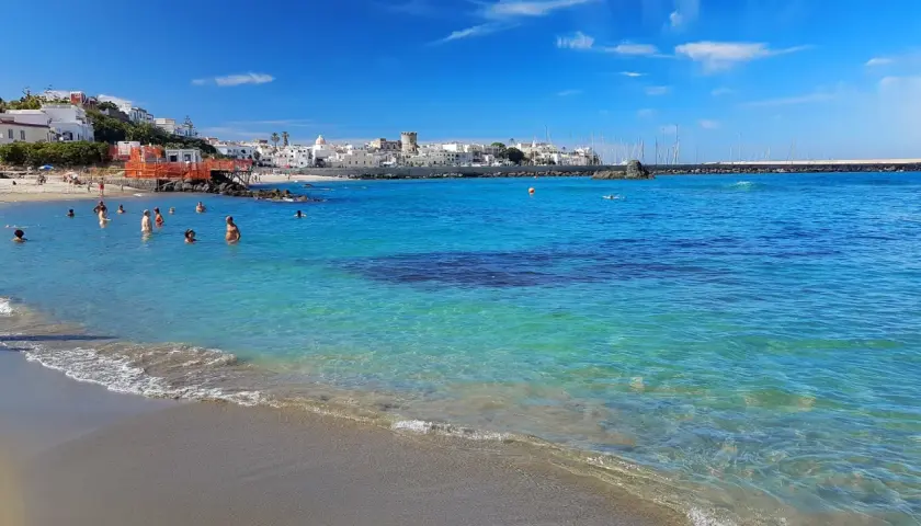 Persone nuotano nelle acque cristalline della spiaggia della Chiaia, con un borgo bianco sullo sfondo e un cielo azzurro con nuvole bianche.