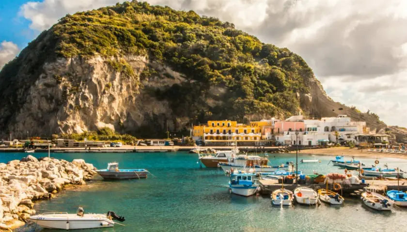Un pittoresco ormeggio a Ischia, con barche colorate ormeggiate davanti a case bianche e gialle, sotto una collina verdeggiante.