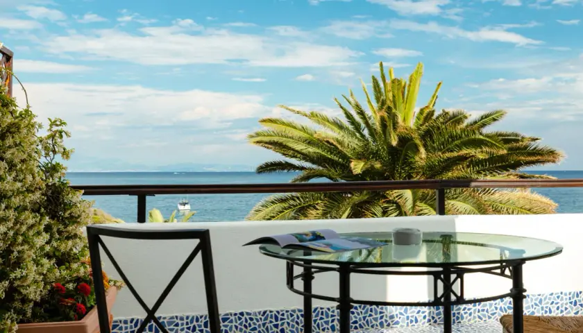 Vista mare da un beach club a Ischia: tavolino in vetro, sedia in ferro e palma rigogliosa sotto un cielo azzurro.
