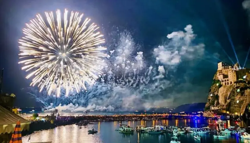 Fuochi d'artificio spettacolari illuminano il cielo notturno sopra un porto pieno di barche, con un castello arroccato su una scogliera. Un'immagine che ricorda le celebrazioni di Mario Mazzella.