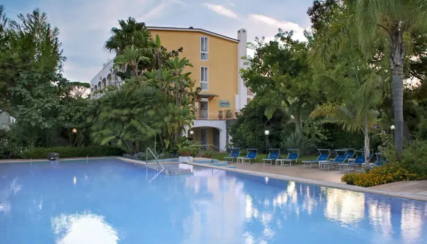 Piscina azzurra e lettini blu in un rigoglioso giardino tropicale, con un edificio giallo sullo sfondo, a San Giovanni, Ischia.