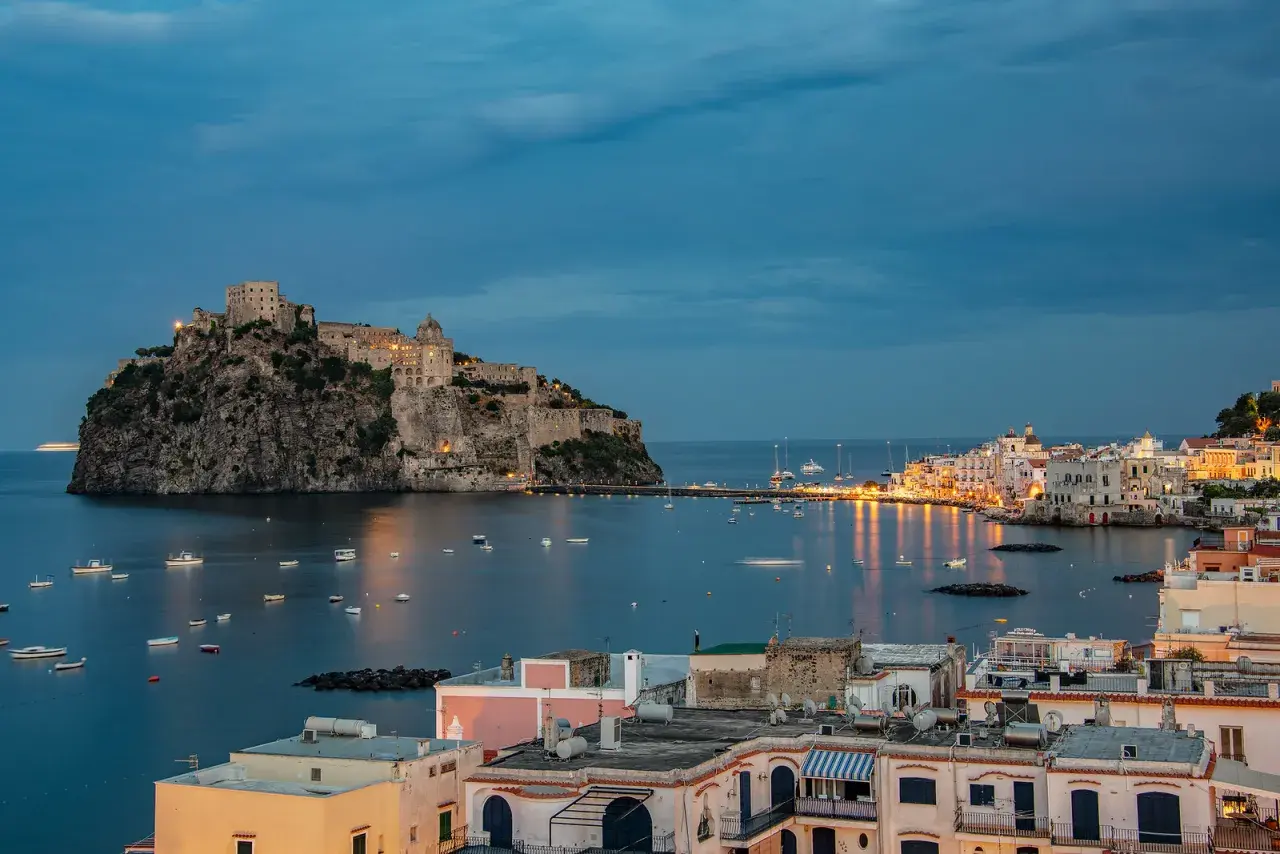 Castello Aragonese domina il mare blu, con barche che ondeggiano e luci che illuminano le pittoresche calette di Ischia al crepuscolo.