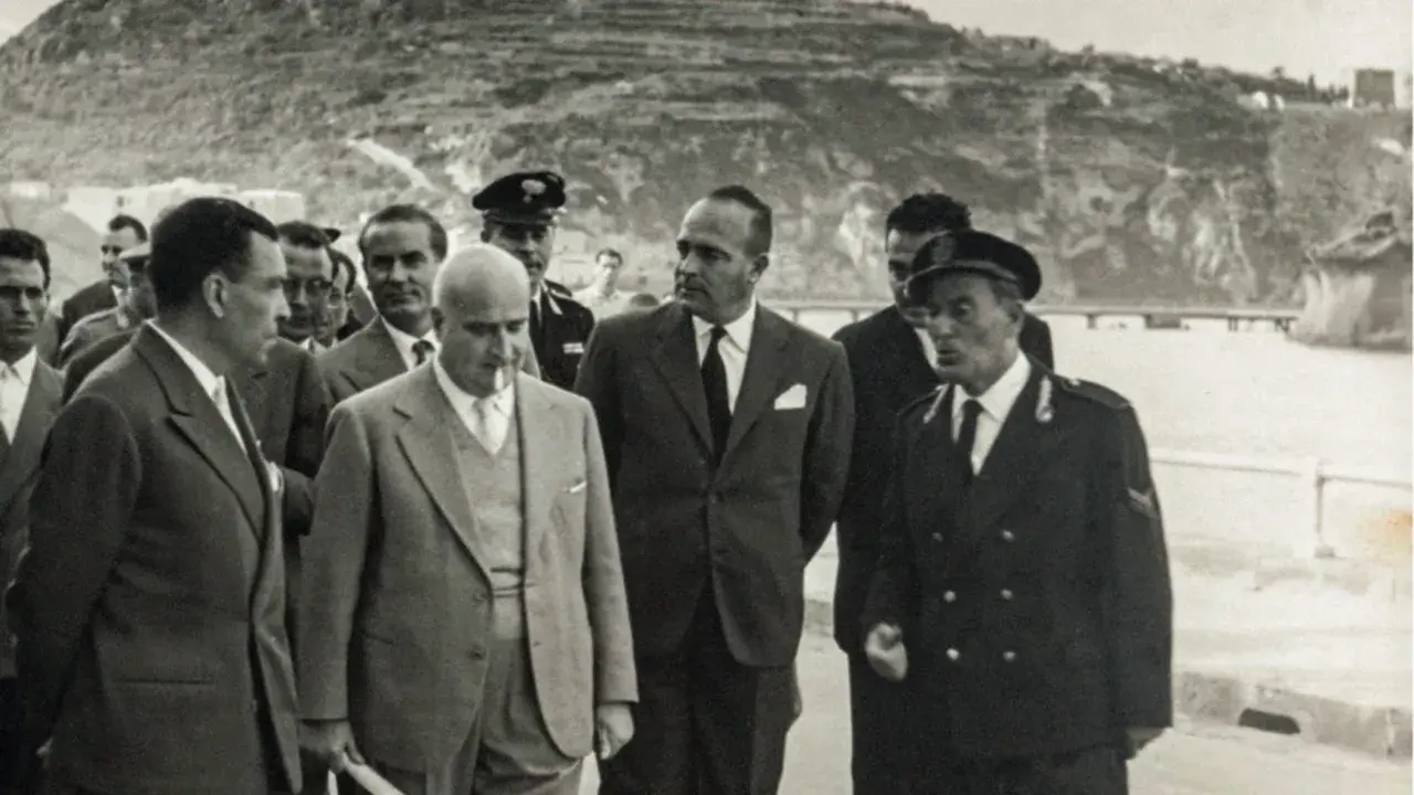 Angelo Rizzoli, con abito scuro, cammina con altri uomini a Ischia, sullo sfondo una collina terrazzata.