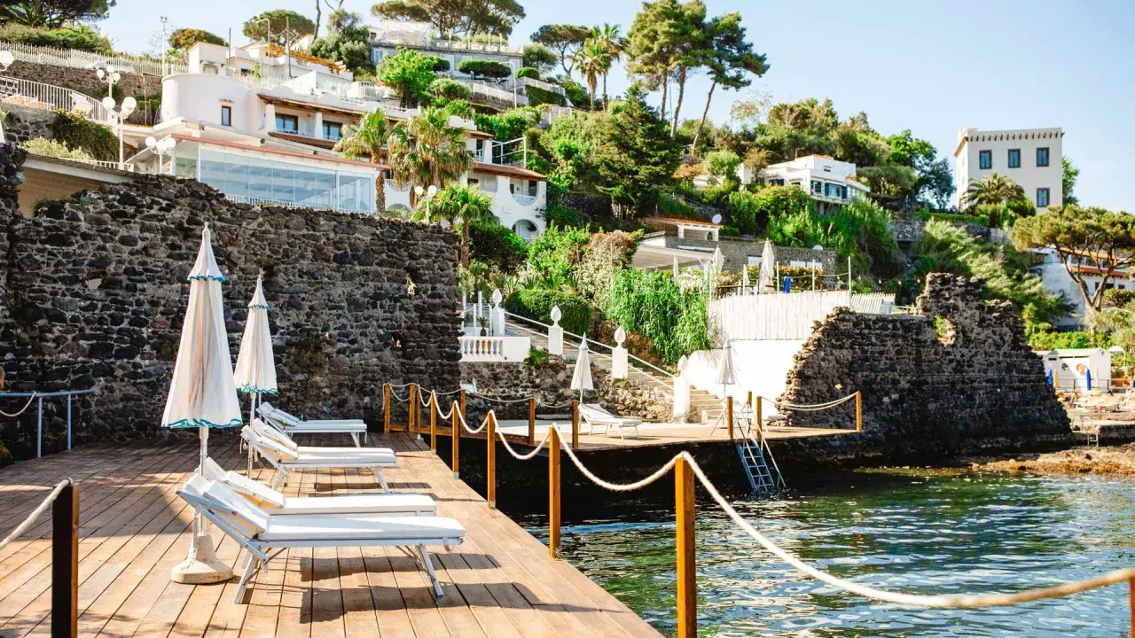 Terrazza in legno con lettini e ombrelloni, vista sul mare e sulle ville del **hotel i delfini Ischia** immerse nel verde.