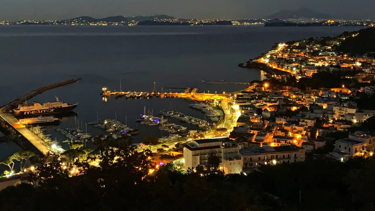 Casamicciola, un gioiello illuminato di notte, con un porto pieno di barche e luci che si riflettono sul mare calmo.