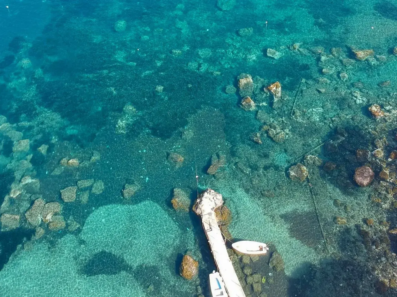 Acque cristalline e fondali rocciosi ad Aenaria, Ischia. Un pontile accoglie piccole imbarcazioni.