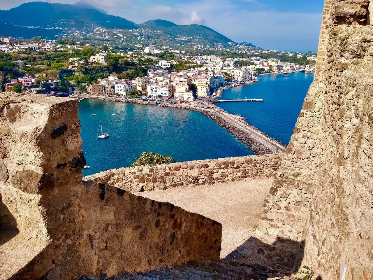 Vista panoramica del porto di Ischia, con i traghetti in attesa e le case colorate che si affacciano sul mare blu.