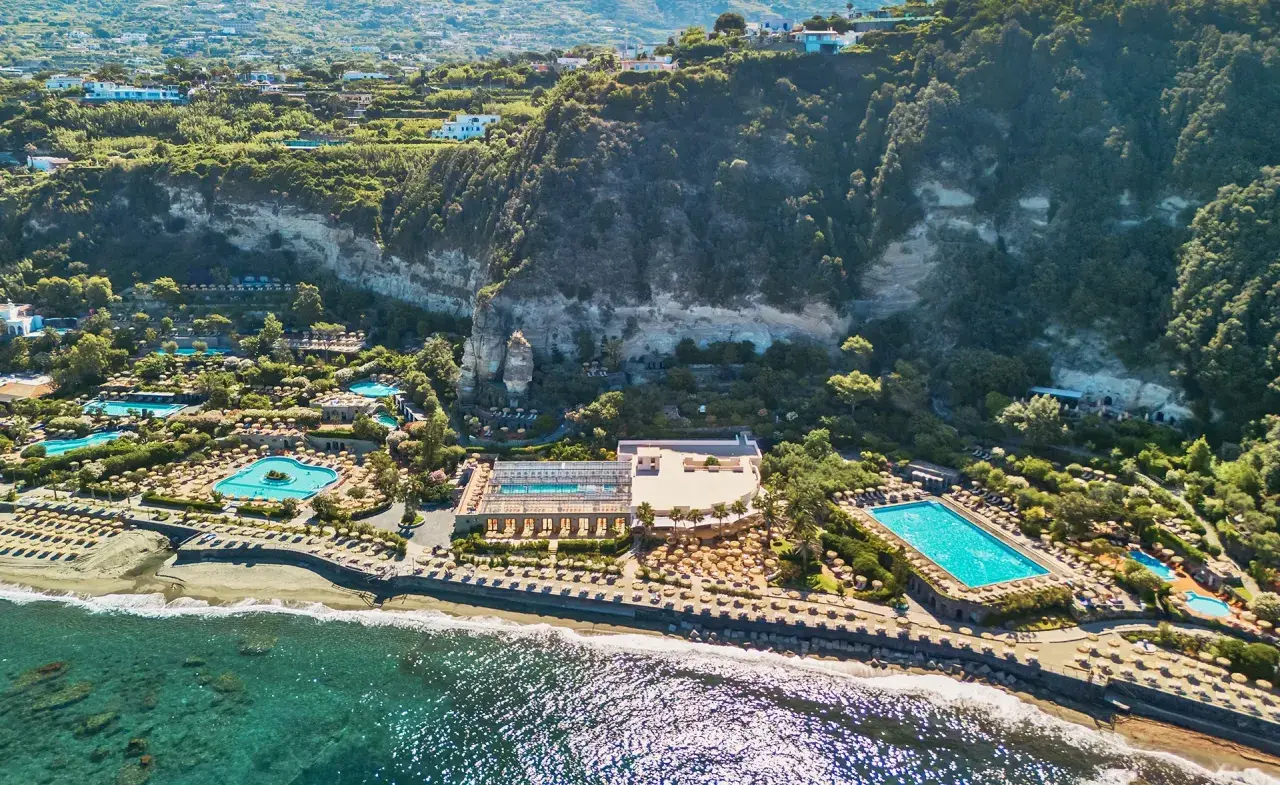 Vista aerea di un resort con piscine e lettini sulla spiaggia, incastonato tra il mare e una scogliera verdeggiante. Un paradiso di terme a Forio.