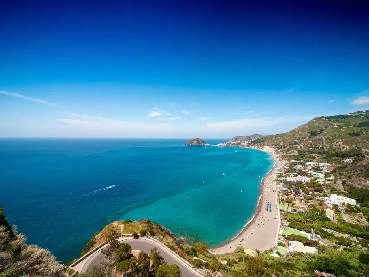 Vista panoramica delle spiagge libere a Ischia, con il mare turchese che lambisce la costa e le case arroccate sulle colline verdi.