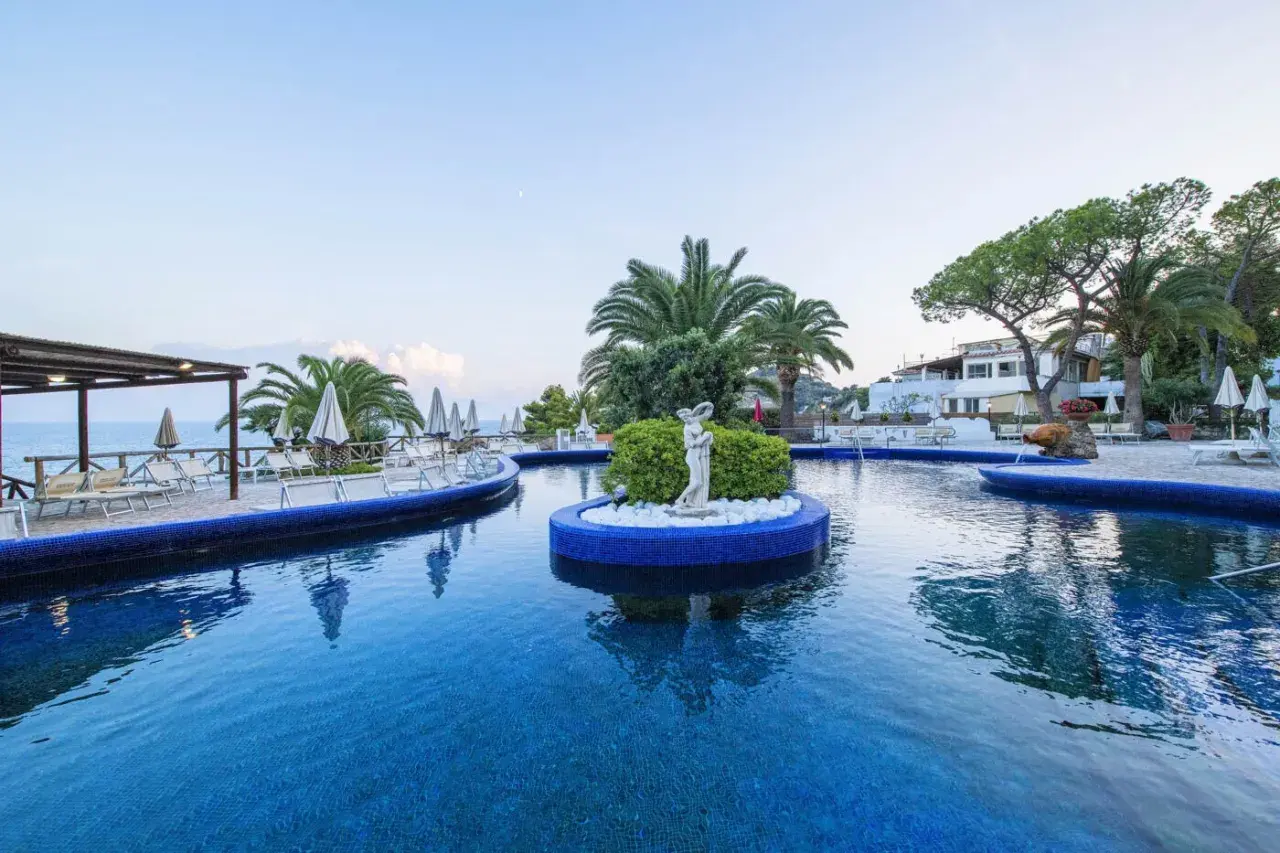 Piscina azzurra con isolotto centrale e statua, lettini e palme intorno. Un'oasi di relax all'Hotel Apollon.