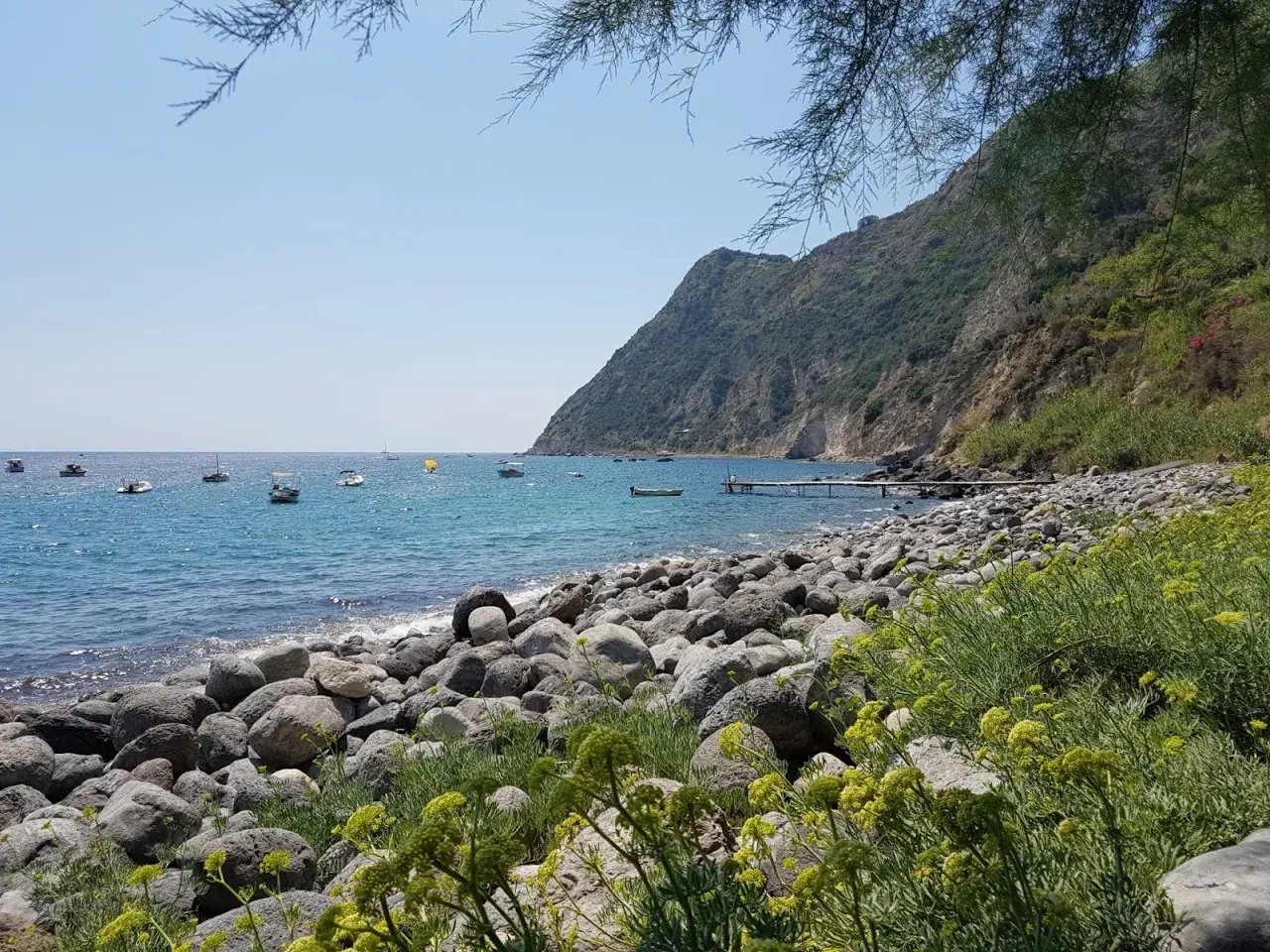 La baia della Scarrupata, con le sue rocce levigate e il mare azzurro, accoglie piccole imbarcazioni. Un pontile si protende nell'acqua.