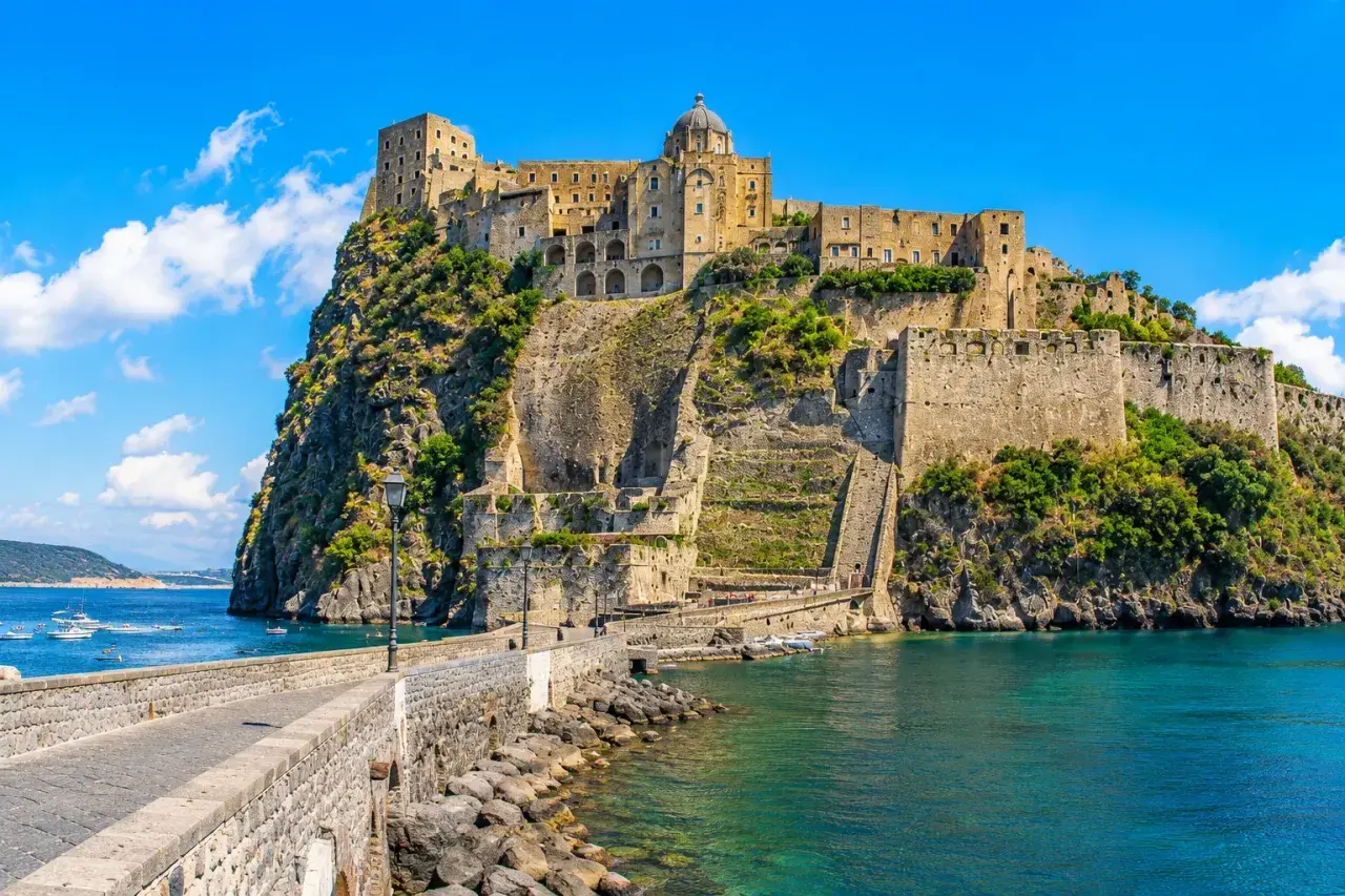 The Aragonese Castle on a rocky island, connected by a stone bridge, with boats in the turquoise sea.