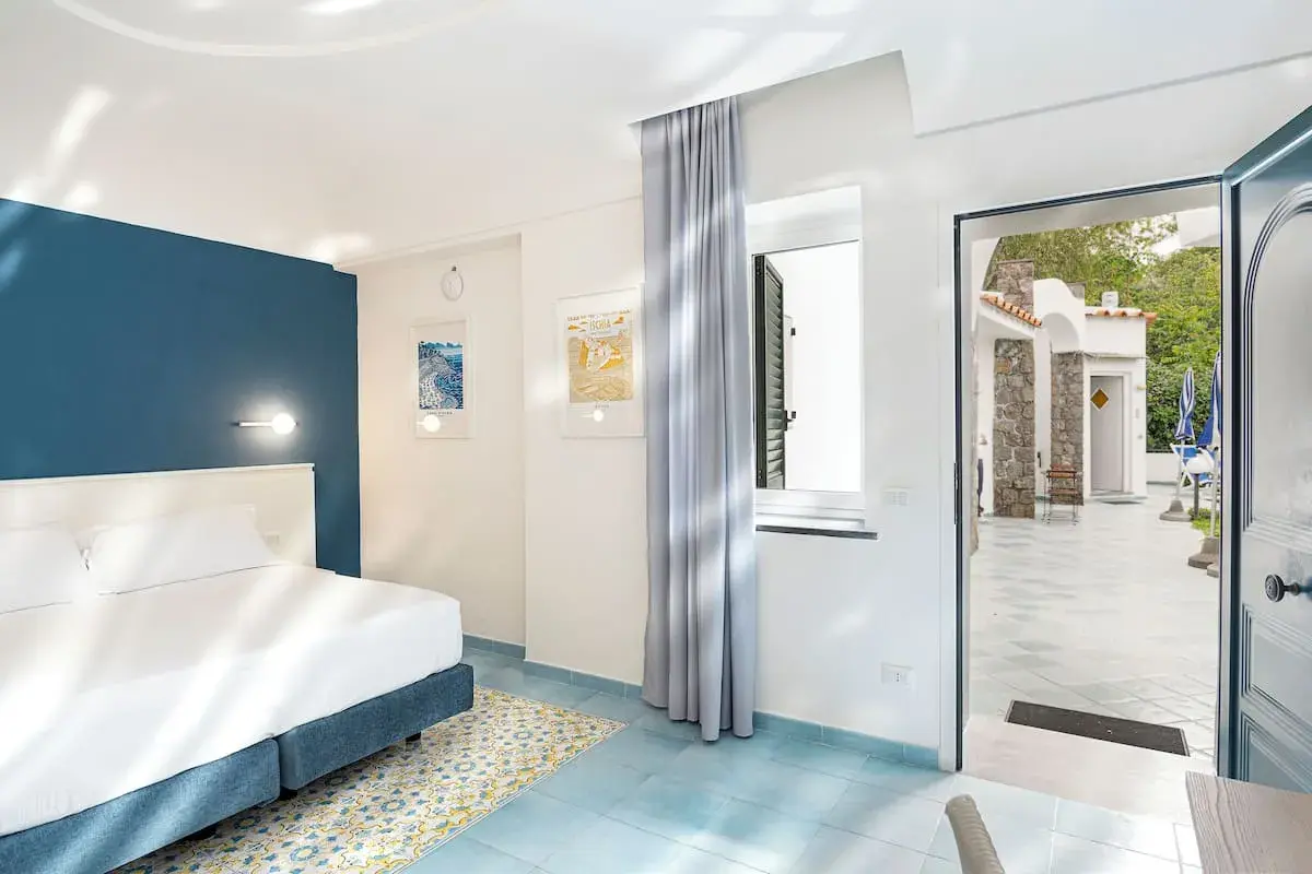 Camera luminosa con letto matrimoniale e vista su un cortile con ombrelloni blu. Un invito a soggiornare all'hotel oasi Ischia.