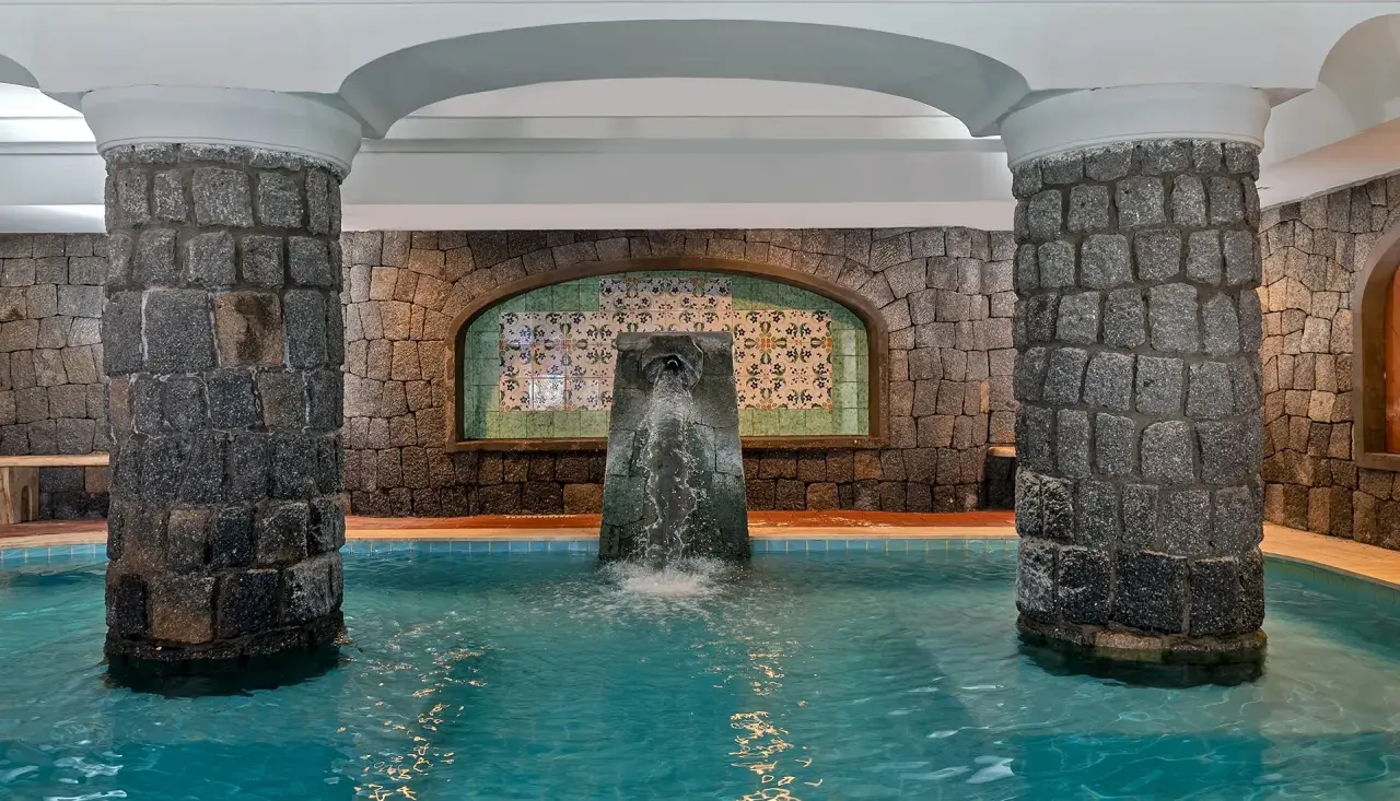 Piscina interna con fontana a cascata, colonne in pietra e piastrelle decorative. Un angolo di relax alle terme di Lacco Ameno.