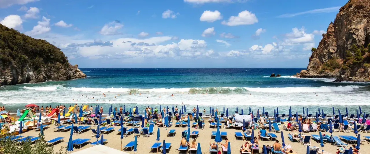 Ischia ad agosto: una spiaggia affollata con lettini blu, ombrelloni e bagnanti che si godono il mare cristallino sotto un cielo azzurro con nuvole bianche.