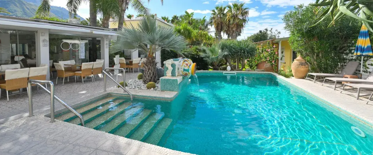 Piscina cristallina con cascata decorativa, lettini prendisole e area ristoro esterna. Un angolo di relax che ricorda le terme comunali di Ischia.