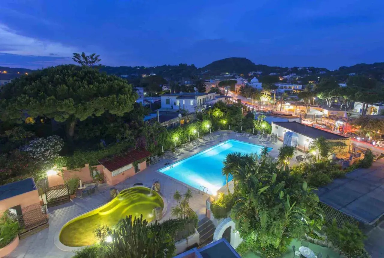 Piscina illuminata e vasca idromassaggio a forma di fagiolo al Hotel Eden Ischia, con vista sul paese di sera.