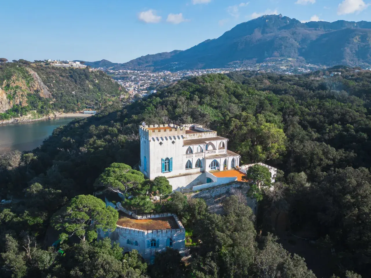 Villa La Colombaia a Ischia, un bianco maniero con torrette e finestre blu, immerso nel verde lussureggiante con vista sul mare e sulle montagne.