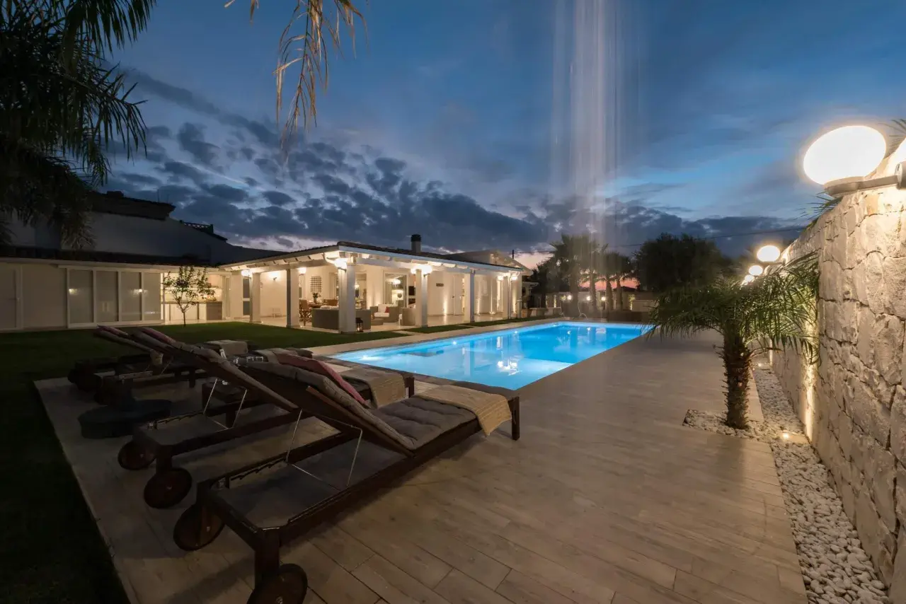 Piscina illuminata e lettini prendisole al tramonto, con cascata d'acqua. Un'oasi di relax al **hotel le palme Ischia**.