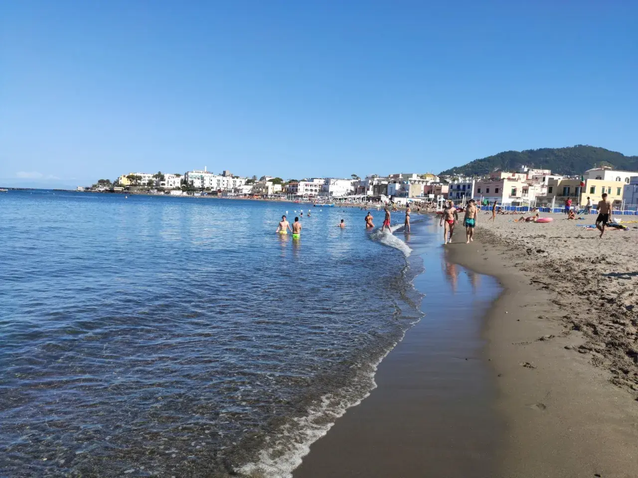 Persone si godono una giornata di sole sulla spiaggia di San Pietro, con il mare azzurro e case colorate sullo sfondo.