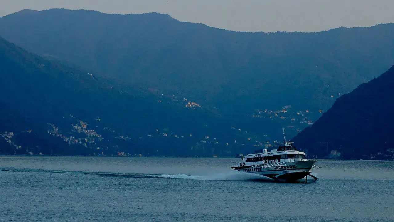 Aliscafo in navigazione su un lago calmo, con montagne boscose e case illuminate sullo sfondo.