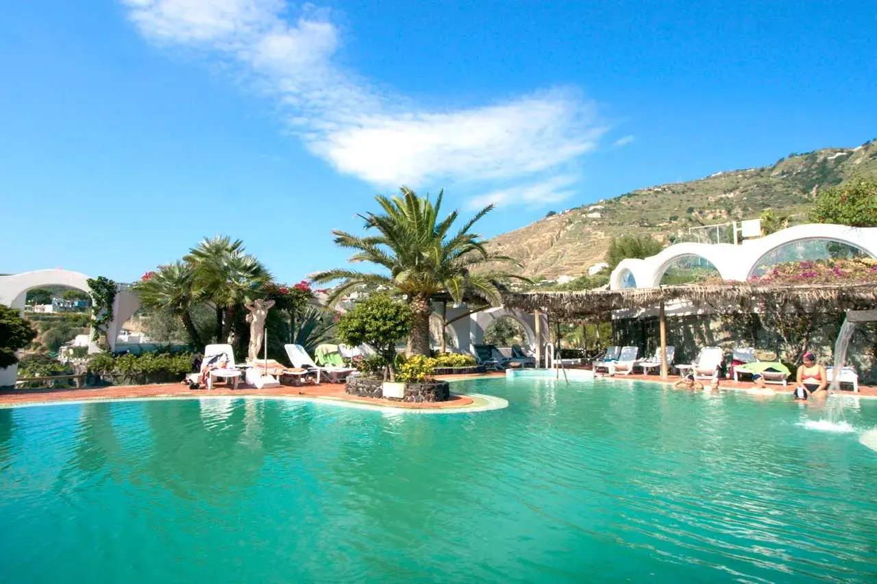 Piscina con lettini e palme, un'oasi di relax alle terme Sant'Angelo Ischia, con vista sulla collina verdeggiante.