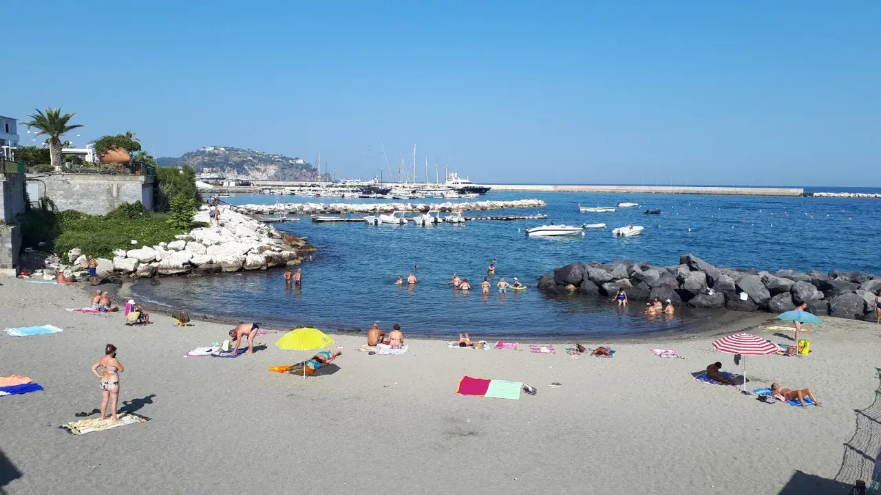 Spiaggia della Marina di Ischia: bagnanti si godono il sole e il mare, con barche ormeggiate nel porto sullo sfondo.