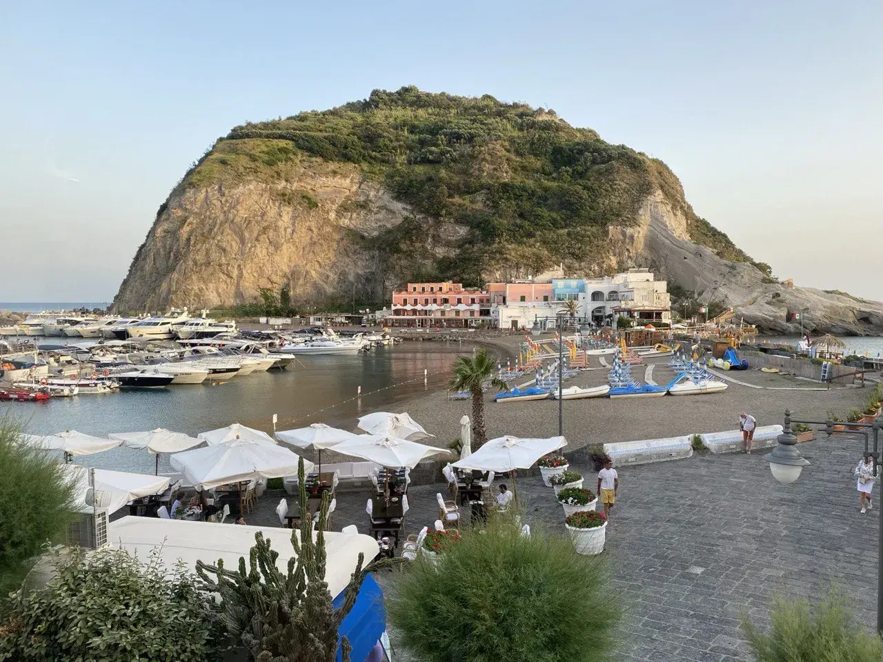 Il porto turistico di Ischia, con yacht ormeggiati, una spiaggia con lettini e ombrelloni, e case colorate ai piedi di una collina rocciosa.