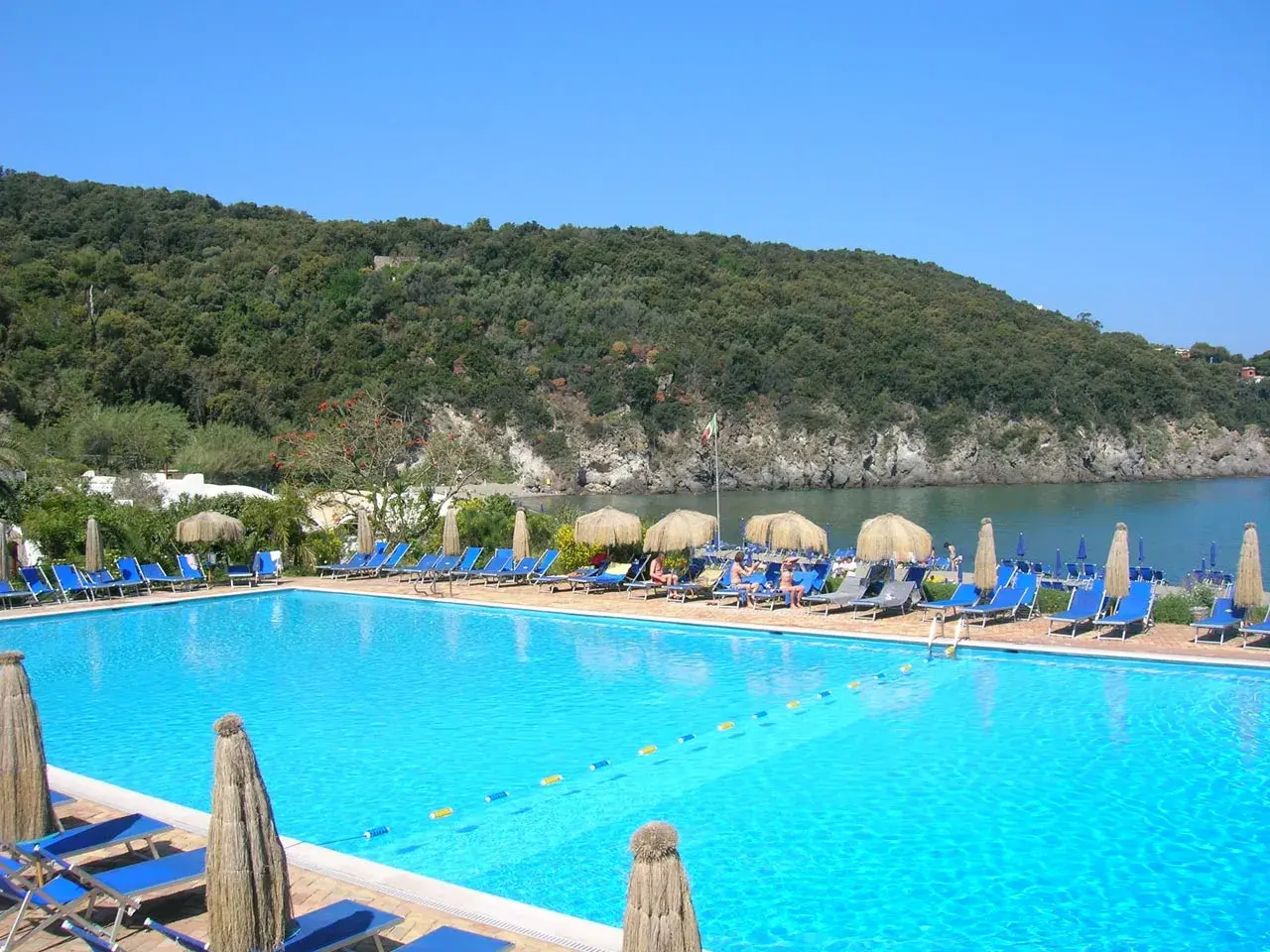 Piscina azzurra con lettini e ombrelloni, vista sul mare e collina verdeggiante. Un angolo di paradiso, forse Negombo o un luogo ispirato a Poseidon.