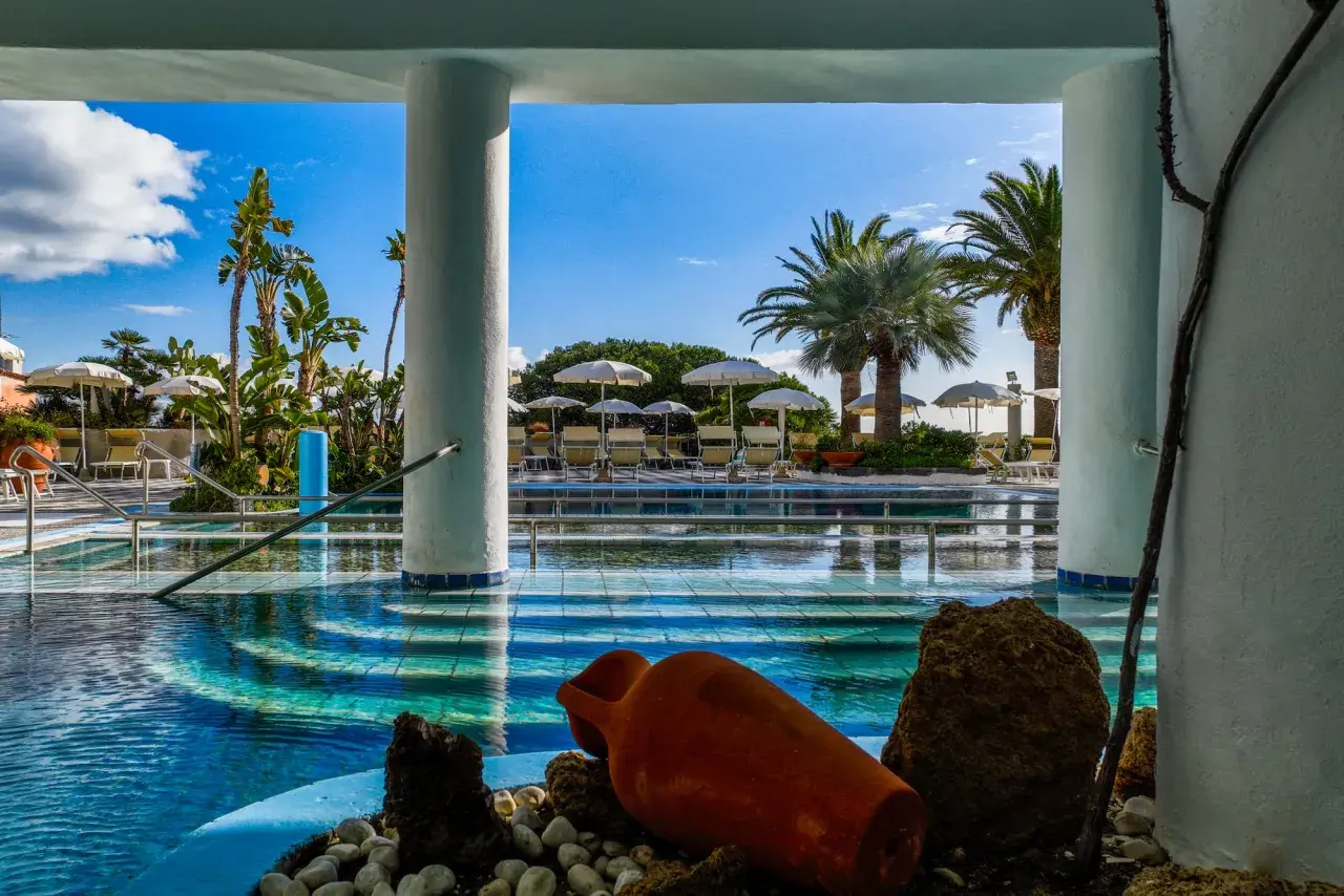Piscina azzurra con palme e lettini, un'atmosfera da **parco termale romantica** con un'antica anfora come decorazione.