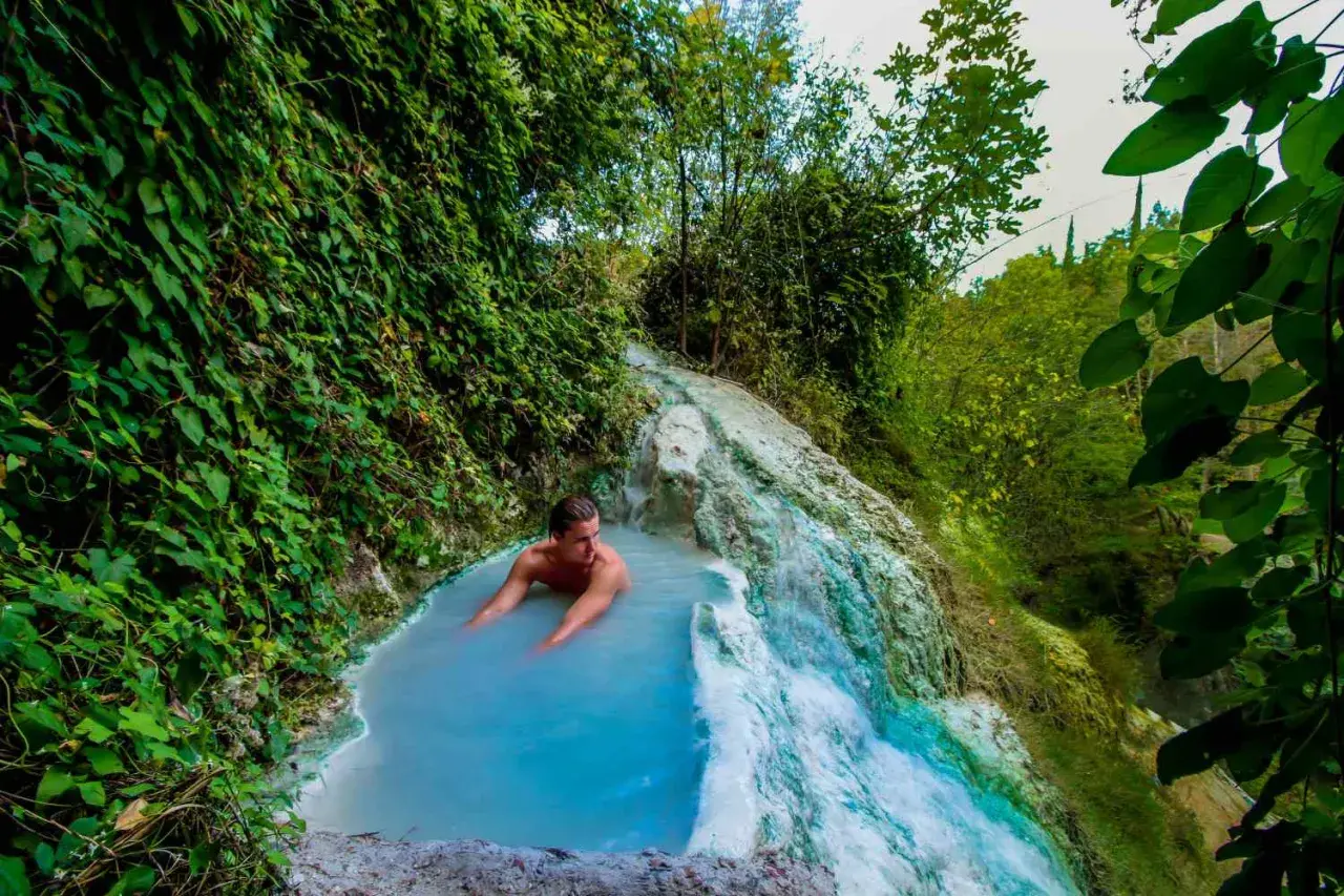 Un uomo si rilassa nelle acque azzurre di una cascata termale naturale a Olmitello, Ischia, circondato da lussureggiante vegetazione.