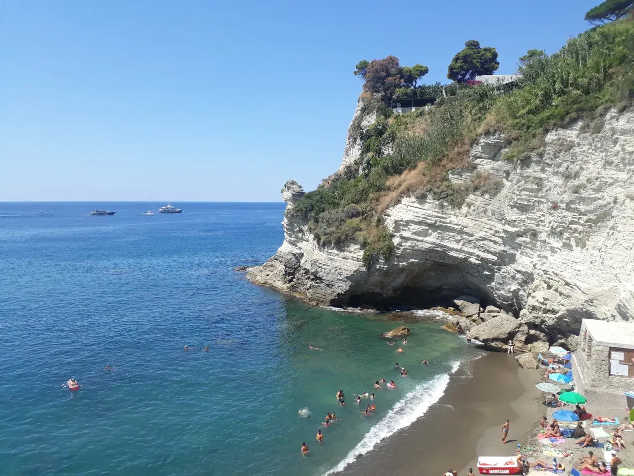 Scogliera bianca a picco sul mare turchese, con bagnanti e yacht all'orizzonte. La **cava grado Ischia** offre un rifugio naturale.